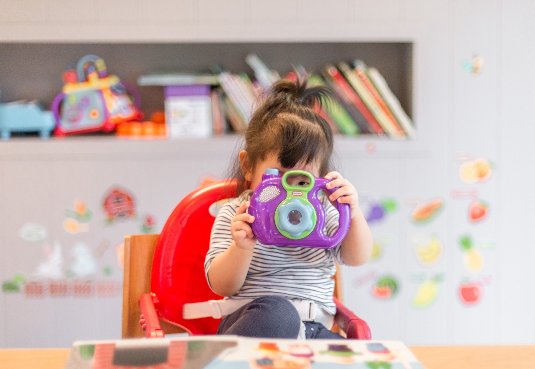 Little girl looks through toy camera lens while reading a picture book and sitting at a table in her homeschool classroom.